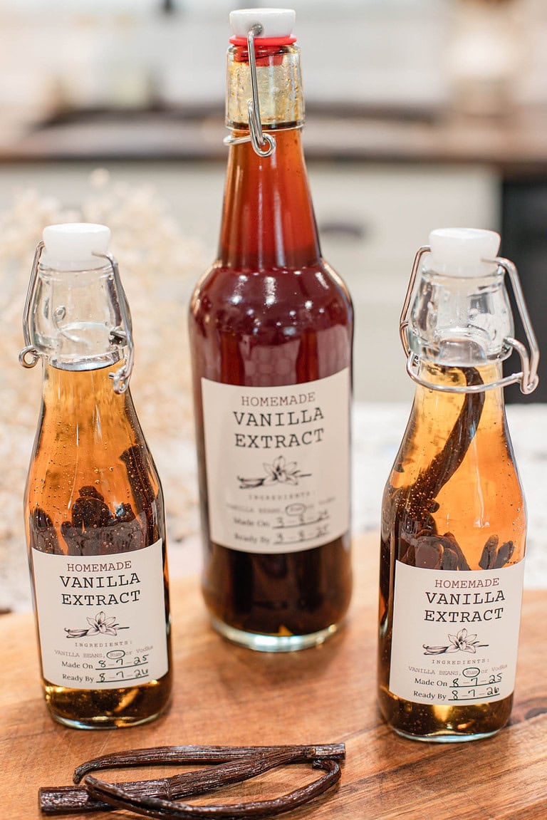 Three swing-top glass bottles of homemade vanilla extract on a wooden board, filled with amber liquid and whole vanilla beans, labeled “Homemade Vanilla Extract.”