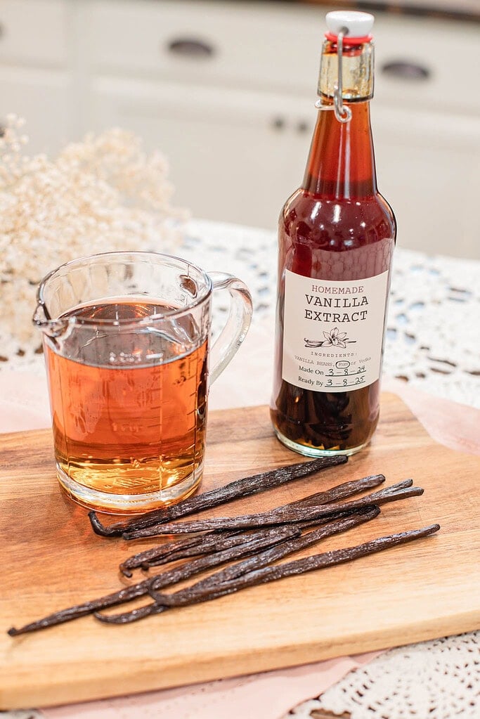 Glass measuring cup filled with homemade vanilla extract beside a labeled bottle and whole vanilla beans on a wooden cutting board.