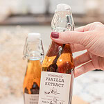 Hand holding a small swing-top bottle of homemade vanilla extract with visible vanilla beans steeping inside.