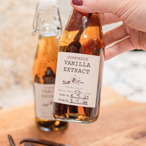 Hand holding a small swing-top bottle of homemade vanilla extract with visible vanilla beans steeping inside.