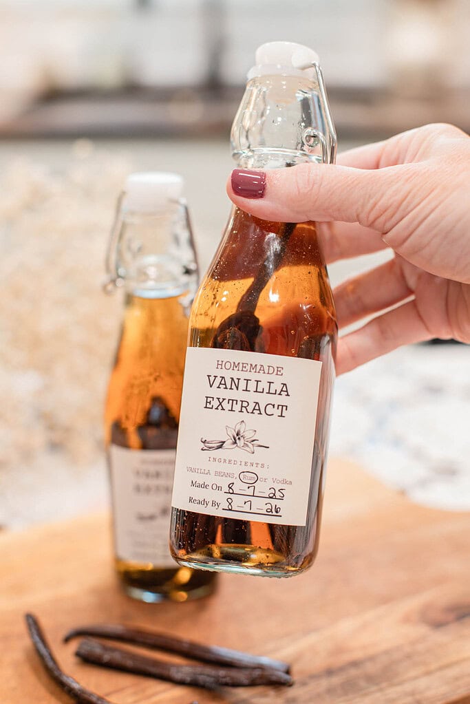 Hand holding a small swing-top bottle of homemade vanilla extract with visible vanilla beans steeping inside.