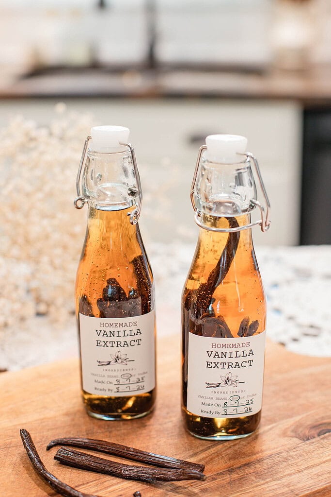 Two small glass bottles of homemade vanilla extract with whole vanilla beans inside, resting on a wooden board with extra beans in front.