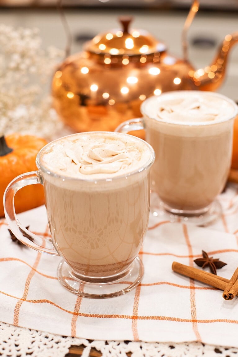 Two pumpkin spice lattes in clear glass mugs topped with whipped cream, styled with a copper teapot, cinnamon sticks, and mini pumpkins