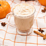 Pumpkin spice latte in a clear mug topped with whipped cream, styled with mini pumpkins, cinnamon sticks, and star anise on a cozy cloth