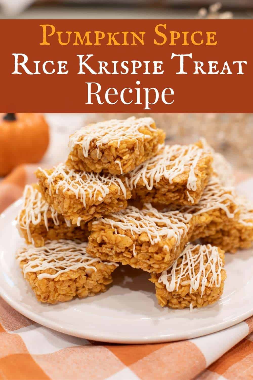 A cozy fall dessert plate stacked with pumpkin spice Rice Krispie treats, golden orange in color and drizzled with white chocolate. Perfect for autumn gatherings, these chewy no-bake bars bring classic marshmallow sweetness with warm pumpkin spice flavor, styled on a white plate over an orange gingham cloth with mini pumpkins in the background.