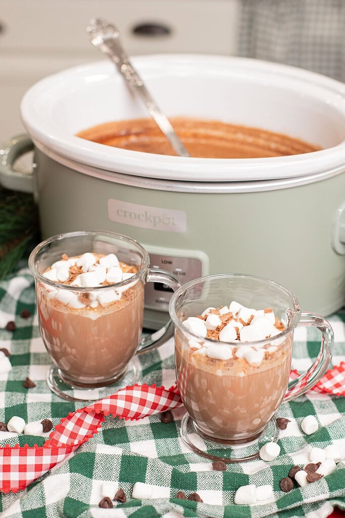 Two glass mugs of creamy crockpot hot chocolate topped with mini marshmallows and chocolate shavings in front of a sage green slow cooker, on a green gingham cloth scattered with chocolate chips and marshmallows