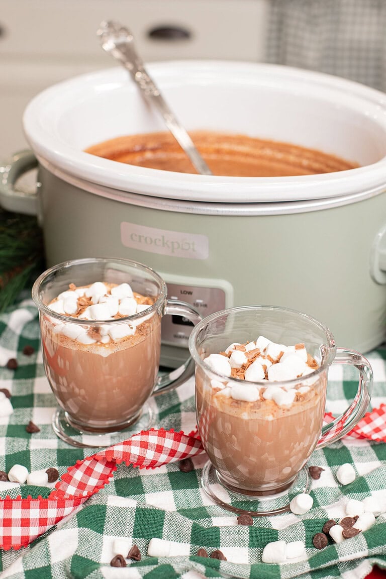 Two glass mugs of creamy crockpot hot chocolate topped with mini marshmallows and chocolate shavings in front of a sage green slow cooker, on a green gingham cloth scattered with chocolate chips and marshmallows