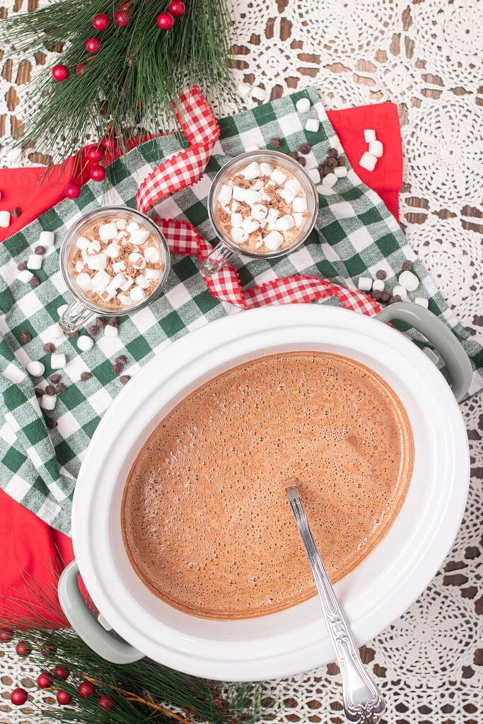 Overhead view of a sage green crockpot filled with steaming hot chocolate, with two glass mugs of hot chocolate topped with mini marshmallows on a green gingham cloth beside pine branches and red berries