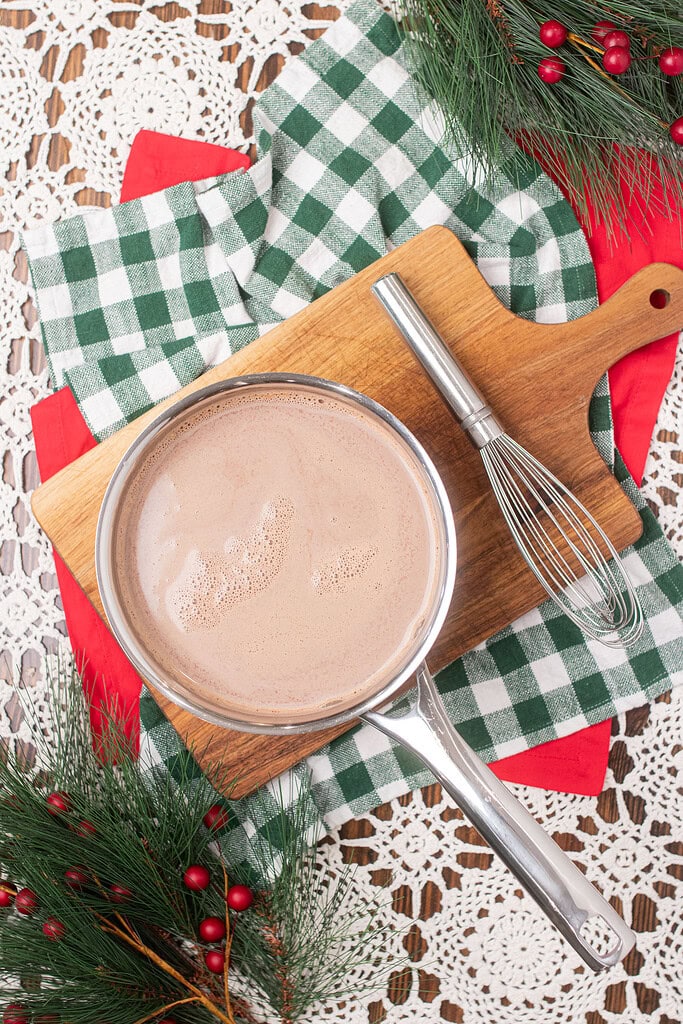 Overhead view of a saucepan of hot chocolate on a wooden cutting board with a whisk, surrounded by a green gingham cloth, pine branches, and red berry sprigs on a lace doily