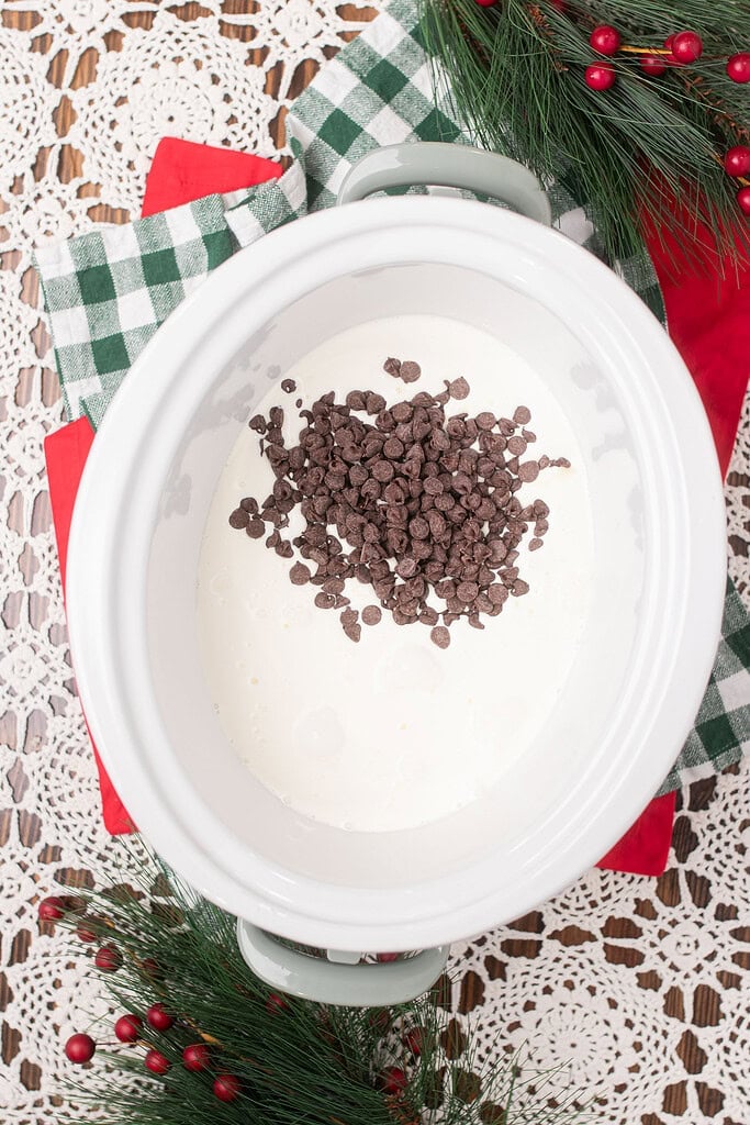 Overhead view of chocolate chips poured into cream inside a white sage green crockpot insert, on a green gingham cloth with pine and red berry branches