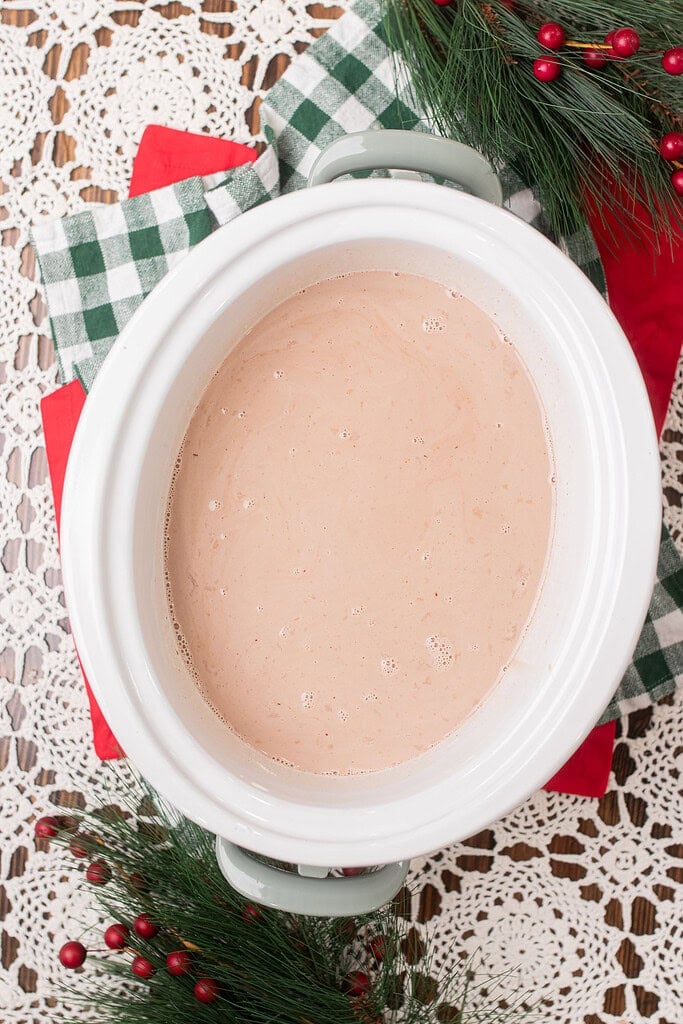 Overhead view of a white crockpot insert filled with lightly stirred hot chocolate mixture in the early stages of slow cooking, surrounded by green gingham cloth and Christmas greenery