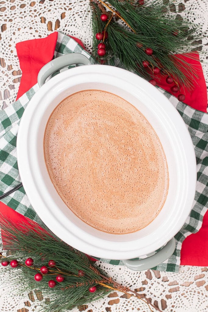 Overhead view of fully heated and frothy crockpot hot chocolate in a white insert set in a sage green slow cooker, surrounded by pine branches, red berries, and a green gingham cloth