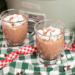 Two glass mugs of creamy crockpot hot chocolate topped with mini marshmallows and chocolate shavings on a green gingham cloth in front of a sage green slow cooker, with scattered chocolate chips and marshmallows