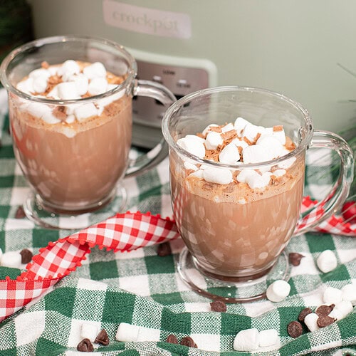 Two glass mugs of creamy crockpot hot chocolate topped with mini marshmallows and chocolate shavings on a green gingham cloth in front of a sage green slow cooker, with scattered chocolate chips and marshmallows