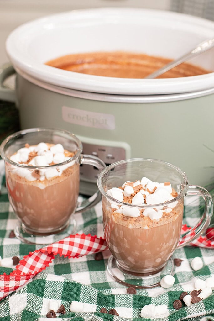 Two glass mugs of creamy crockpot hot chocolate topped with mini marshmallows and chocolate shavings on a green gingham cloth in front of a sage green slow cooker, with scattered chocolate chips and marshmallows