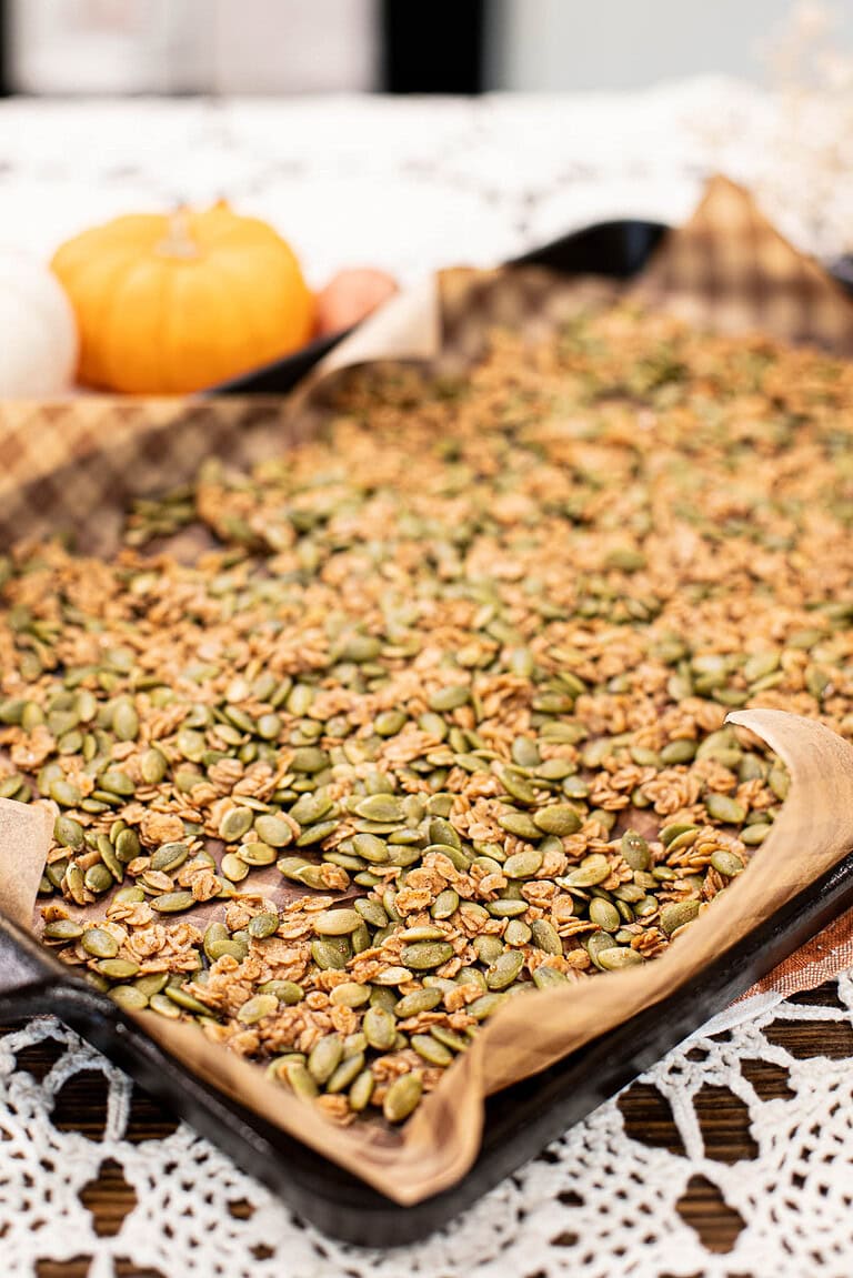 Pumpkin seed brittle spread evenly on a parchment-lined baking sheet with oats and pepitas, with small pumpkins in the background