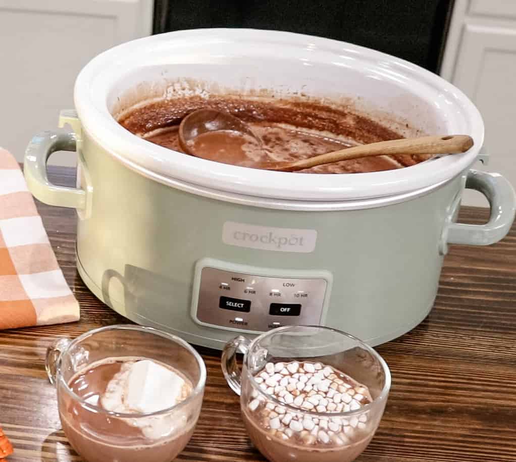 A green crock pot with hot chocolate with two mugs in front of the v=crockpot with hot chocolate and marshmallows.