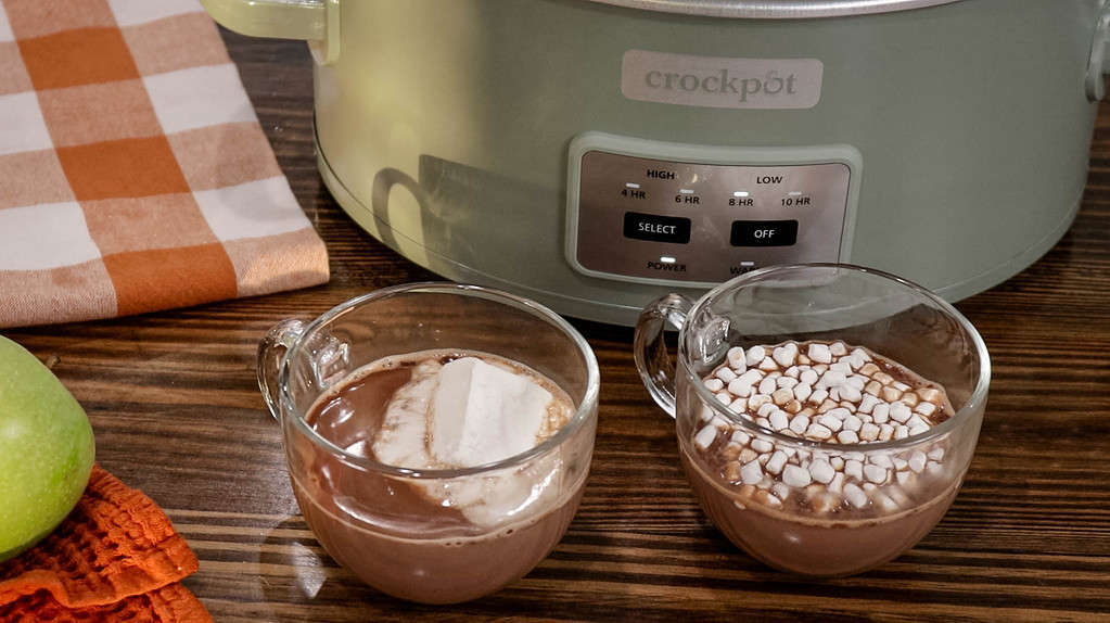 Hot Chocolate in two clear glass mugs with marshmallows in front of a crock pot.