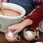 two women in a kitch wearing aprons standing behind a green crock pot one is placing a marshmallow into a cup of hot chocolate.