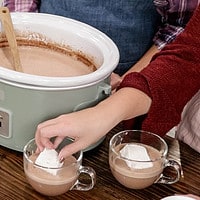 two women in a kitch wearing aprons standing behind a green crock pot one is placing a marshmallow into a cup of hot chocolate.