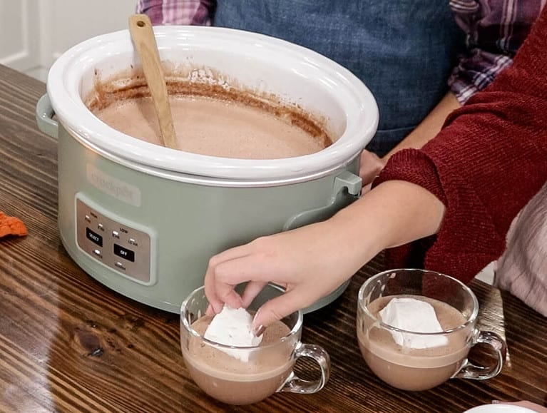 two women in a kitch wearing aprons standing behind a green crock pot one is placing a marshmallow into a cup of hot chocolate.