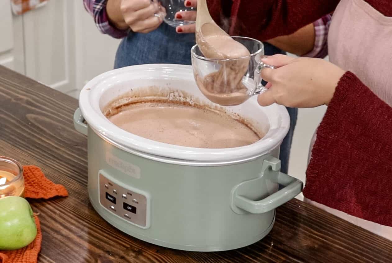 A woman in a pink apron is ladling in crock pot hot chocolate into her mug with a wooden ladle.