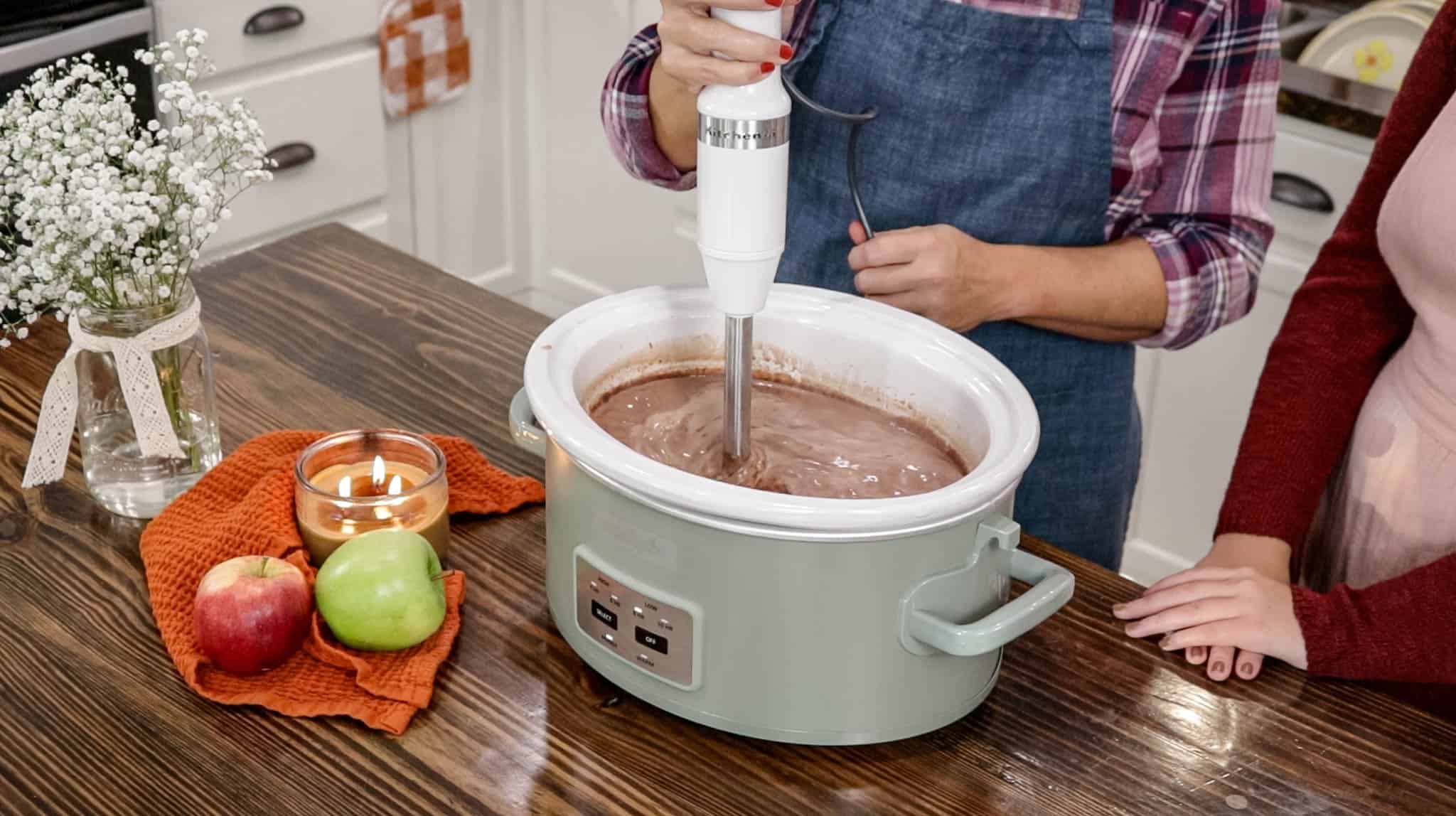 A woman in a blue apron is using a kitchen aid immersion blender in a crock pot mixing up hot chocolate.