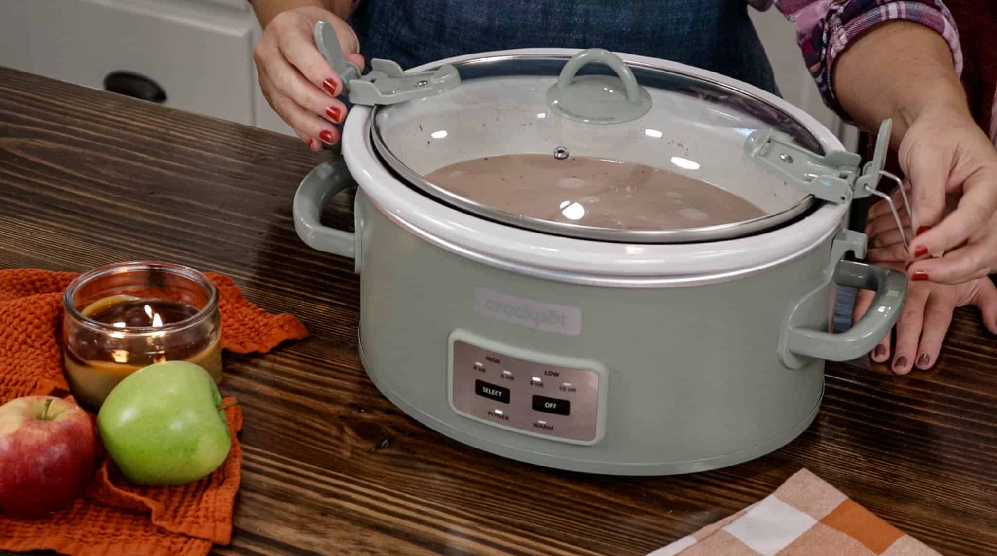 A woman in a apron is putting on the top for a crockpot to cook hot chocolate.