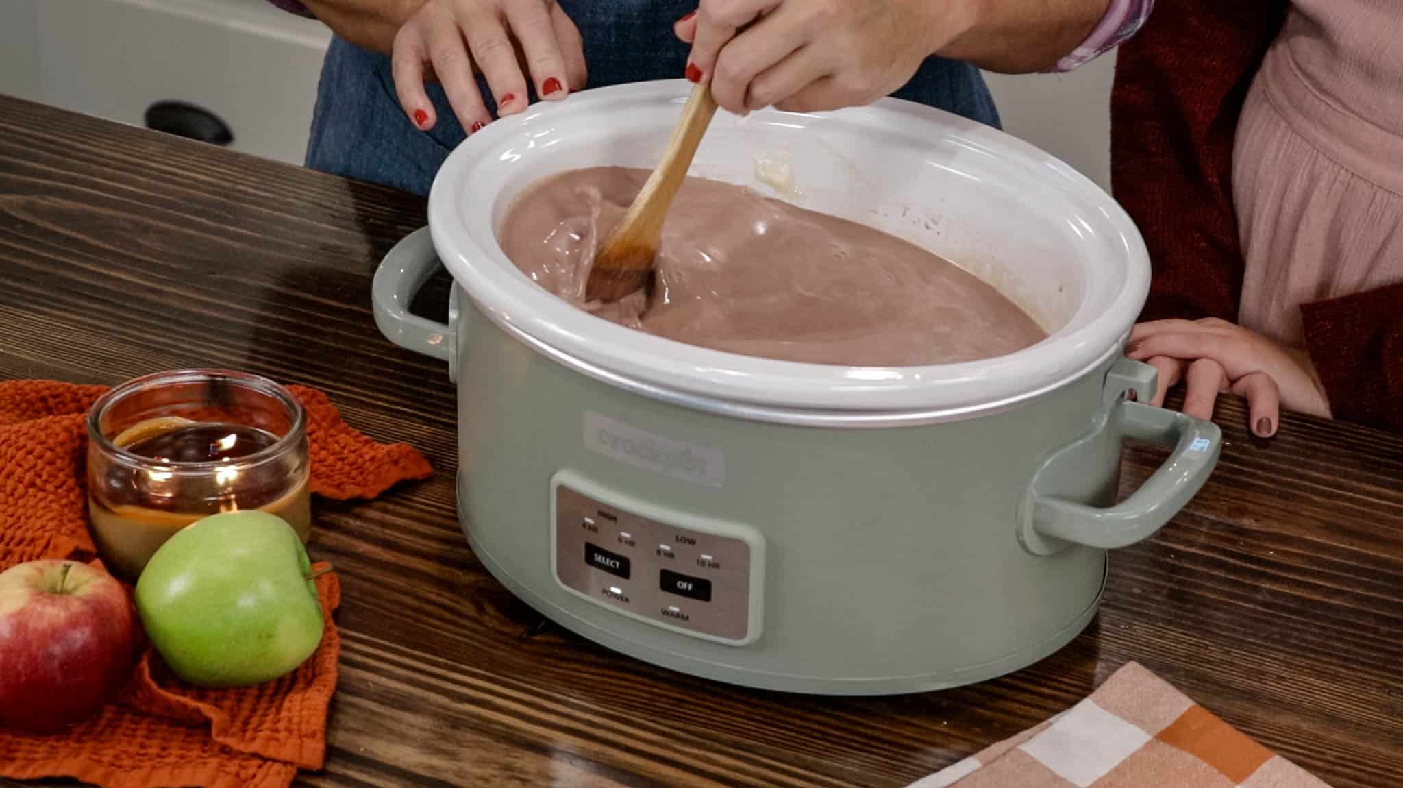 A woman is mixing up hot chocolate in a sage green crock pot with a wooden spoon.
