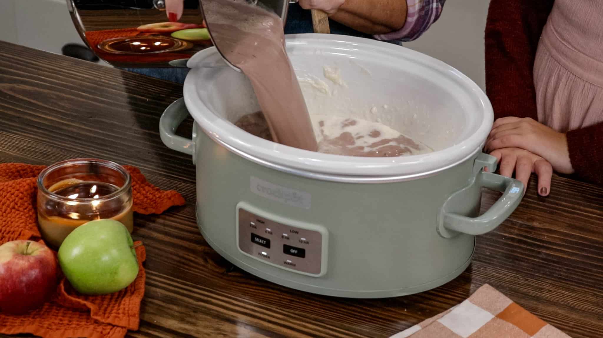 A woman in an apron is pouring in cocoa milk mixture into a crock pot.
