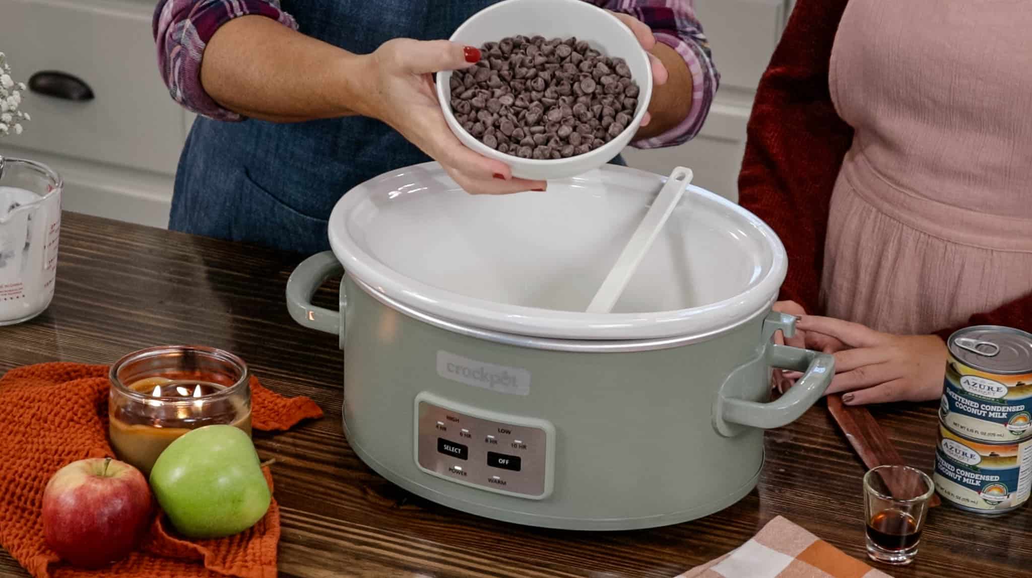 Two woman in a kitchen one is holding a bowl full of chocolate chips over a sage green crockpot.