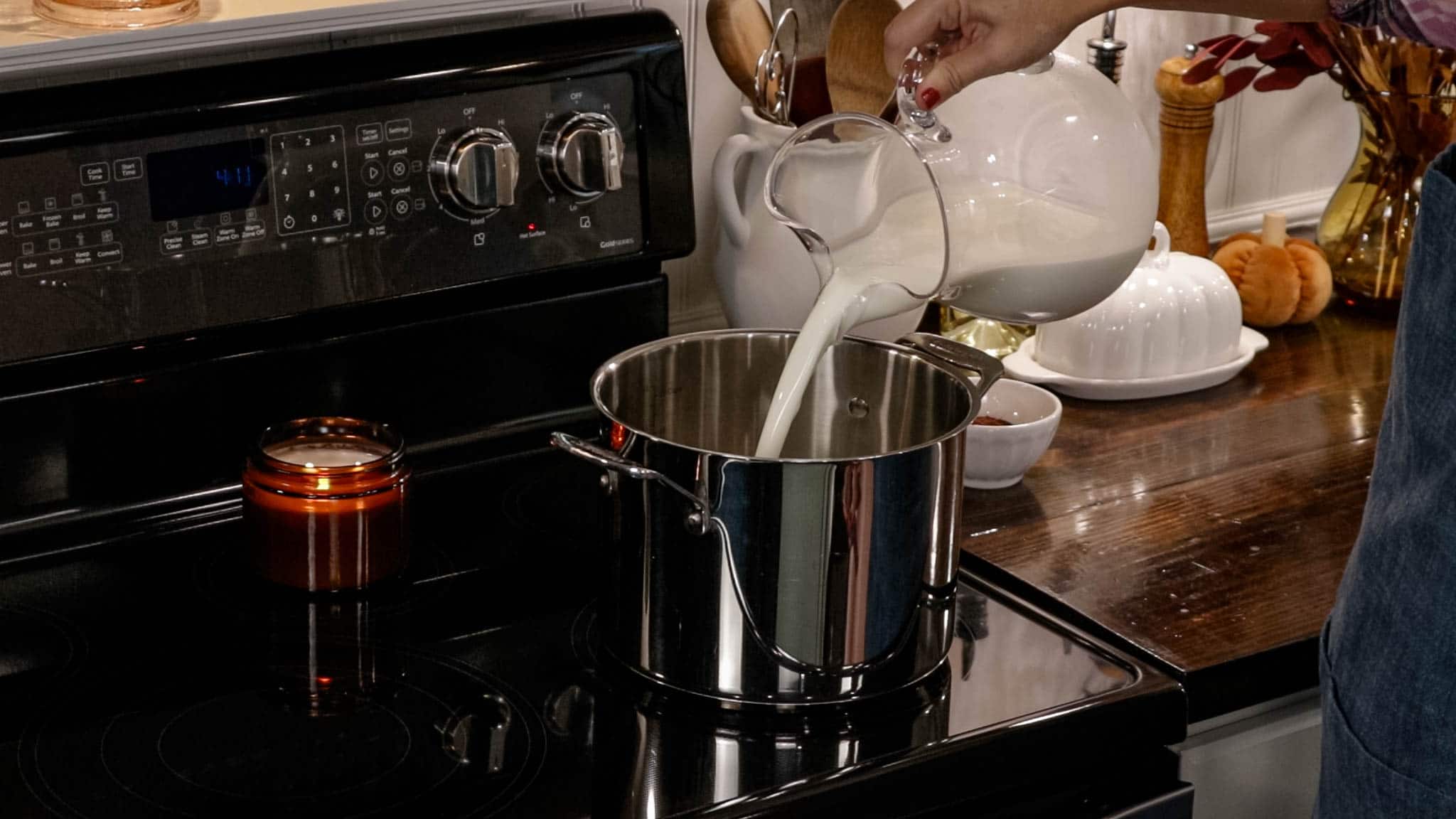 A woman in a blue apron is pouring in whole milk into a stock pot using a clear glass pitcher.