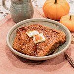 A slice of sourdough pumpkin French toast topped with melting butter pats and maple syrup served on a handmade ceramic plate, styled with mini pumpkins and a stoneware creamer on a dusty rose linen.