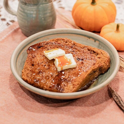 A slice of sourdough pumpkin French toast topped with melting butter pats and maple syrup served on a handmade ceramic plate, styled with mini pumpkins and a stoneware creamer on a dusty rose linen.