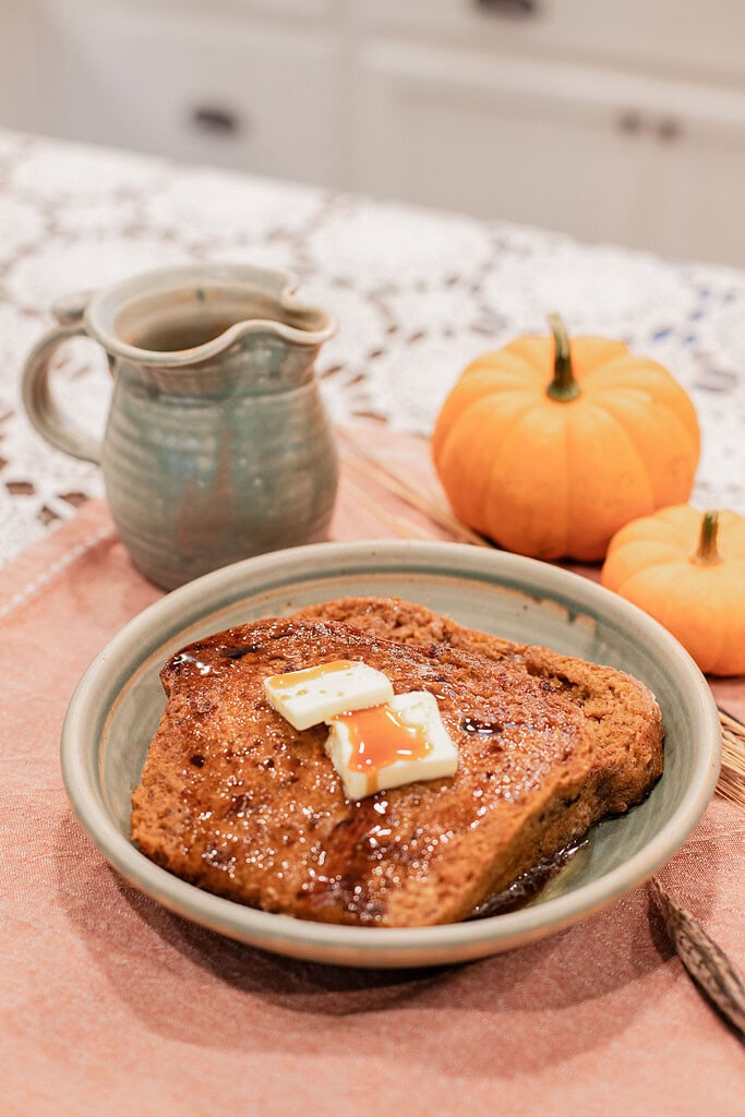 A slice of sourdough pumpkin French toast topped with melting butter pats and maple syrup served on a handmade ceramic plate, styled with mini pumpkins and a stoneware creamer on a dusty rose linen.