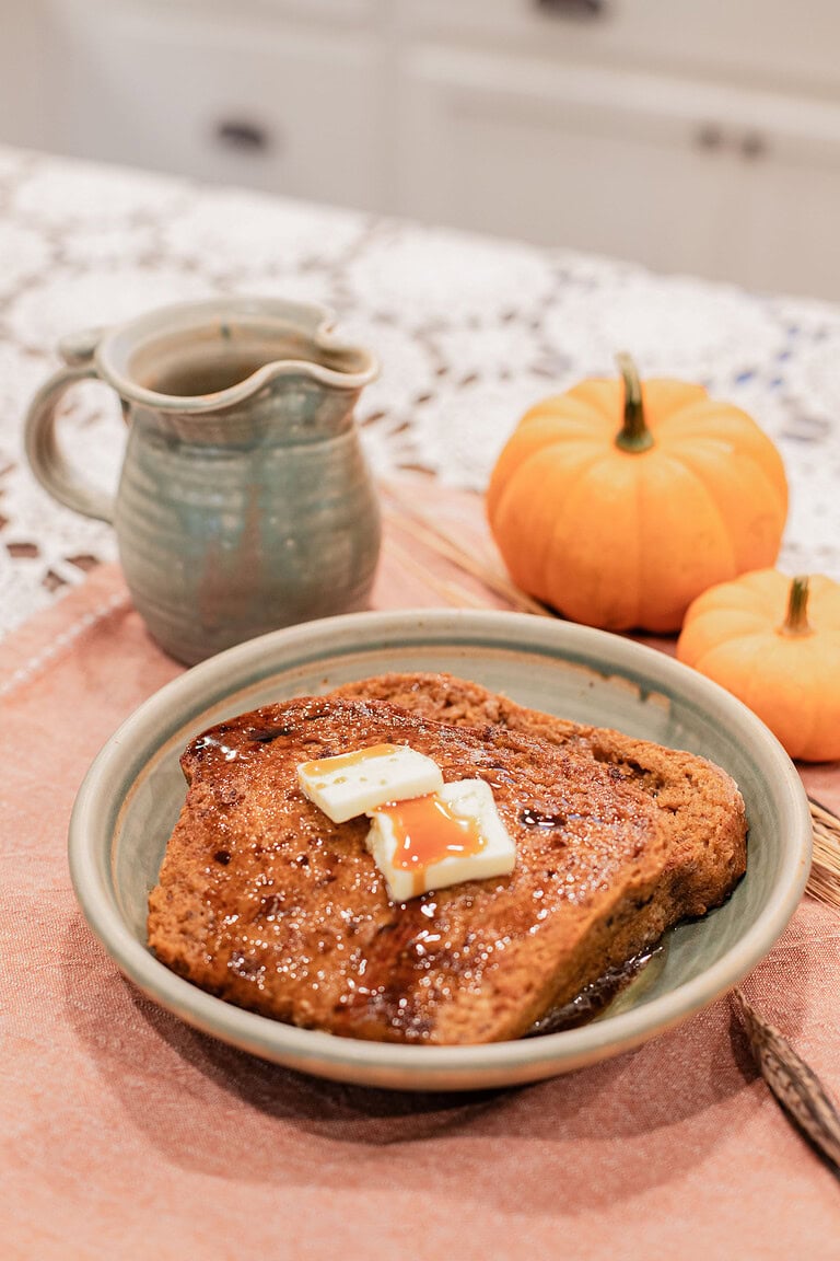 A slice of sourdough pumpkin French toast topped with melting butter pats and maple syrup served on a handmade ceramic plate, styled with mini pumpkins and a stoneware creamer on a dusty rose linen.