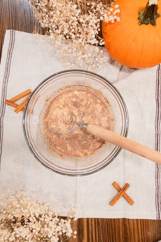 Overhead view of a glass mixing bowl with pumpkin French toast custard batter being whisked, surrounded by cinnamon sticks, dried baby's breath, and a large orange pumpkin on a striped linen towel.