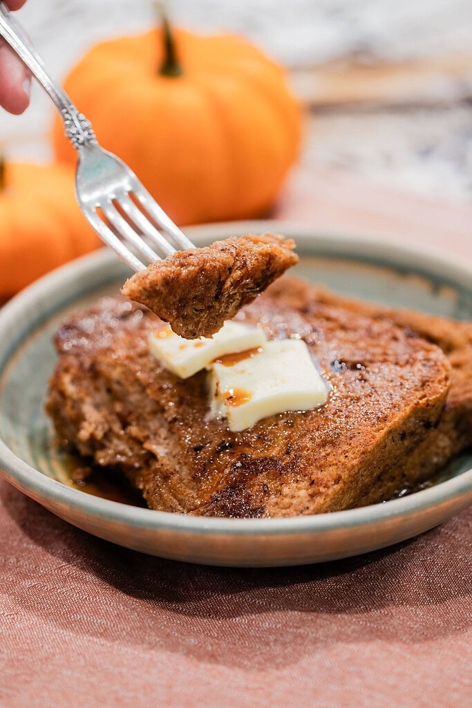 A fork lifting a bite of golden sourdough pumpkin French toast from a ceramic plate topped with butter pats and maple syrup, with mini pumpkins softly blurred in the background.