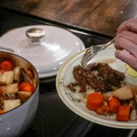 A woman is making a plate of roast beef from a dutch oven on top of a stove.