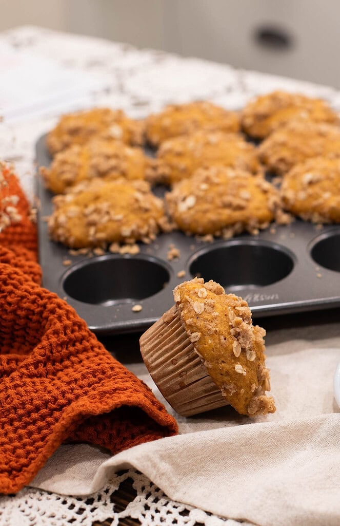 Sourdough pumpkin muffins with oat streusel topping in muffin tin with one muffin leaning in foreground