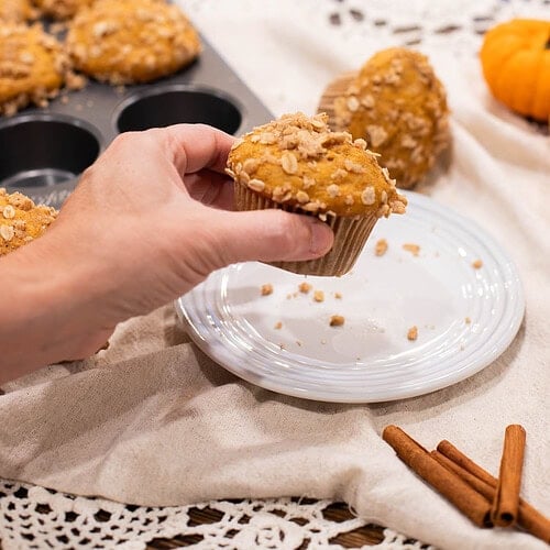 Hand holding sourdough pumpkin muffin with oat streusel topping over plate and kitchen cloth