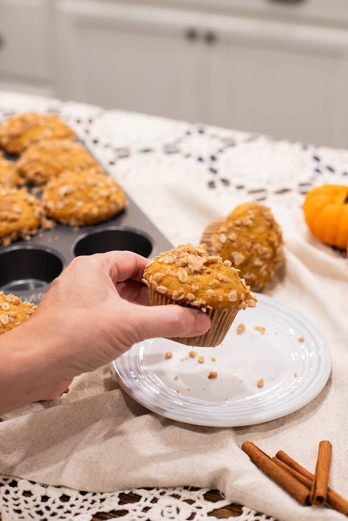 Hand holding sourdough pumpkin muffin with oat streusel topping over plate and kitchen cloth