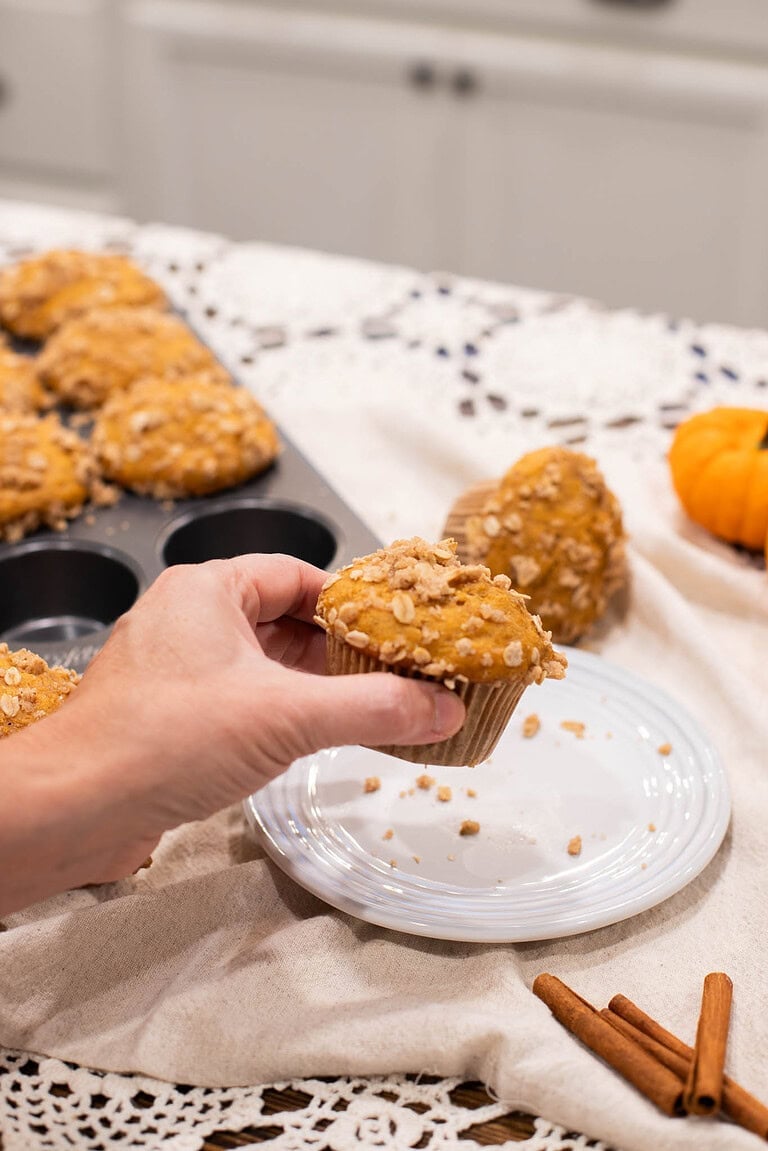 Hand holding sourdough pumpkin muffin with oat streusel topping over plate and kitchen cloth
