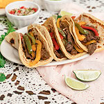 Sourdough tortilla tacos filled with beef fajita meat, peppers, and onions, plated with lime wedges and fresh toppings in a cozy kitchen setting.