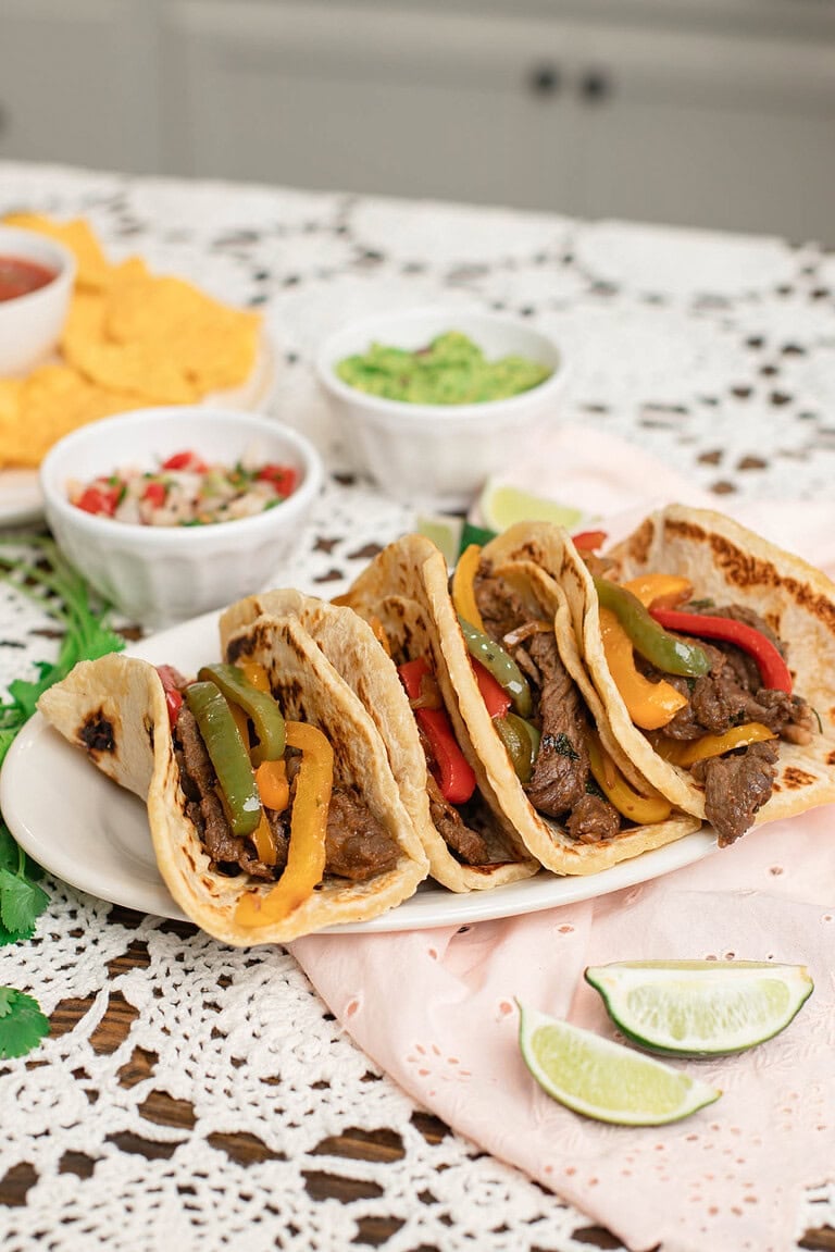 Sourdough tortilla tacos filled with beef fajita meat, peppers, and onions, plated with lime wedges and fresh toppings in a cozy kitchen setting.
