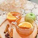 wo glass mugs of hot Dutch oven apple cider garnished with orange slices on a wood slice board, with cinnamon sticks, star anise, and a green apple beside them on a lace doily