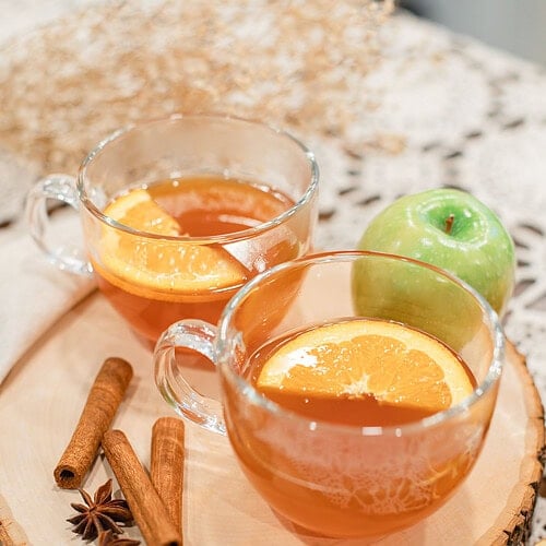 wo glass mugs of hot Dutch oven apple cider garnished with orange slices on a wood slice board, with cinnamon sticks, star anise, and a green apple beside them on a lace doily