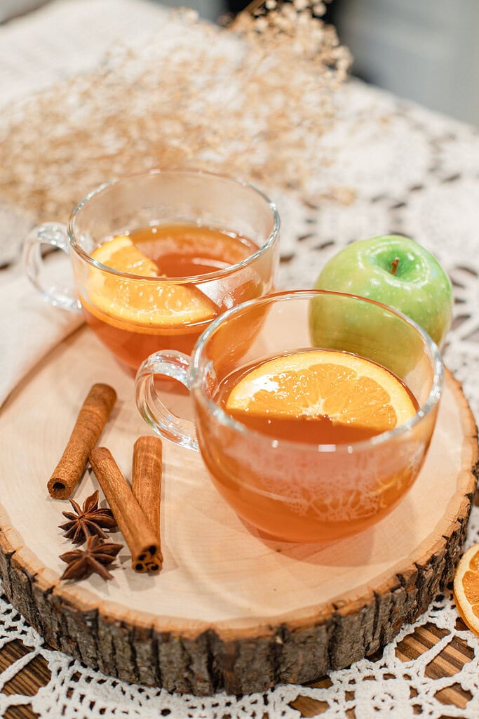 wo glass mugs of hot Dutch oven apple cider garnished with orange slices on a wood slice board, with cinnamon sticks, star anise, and a green apple beside them on a lace doily