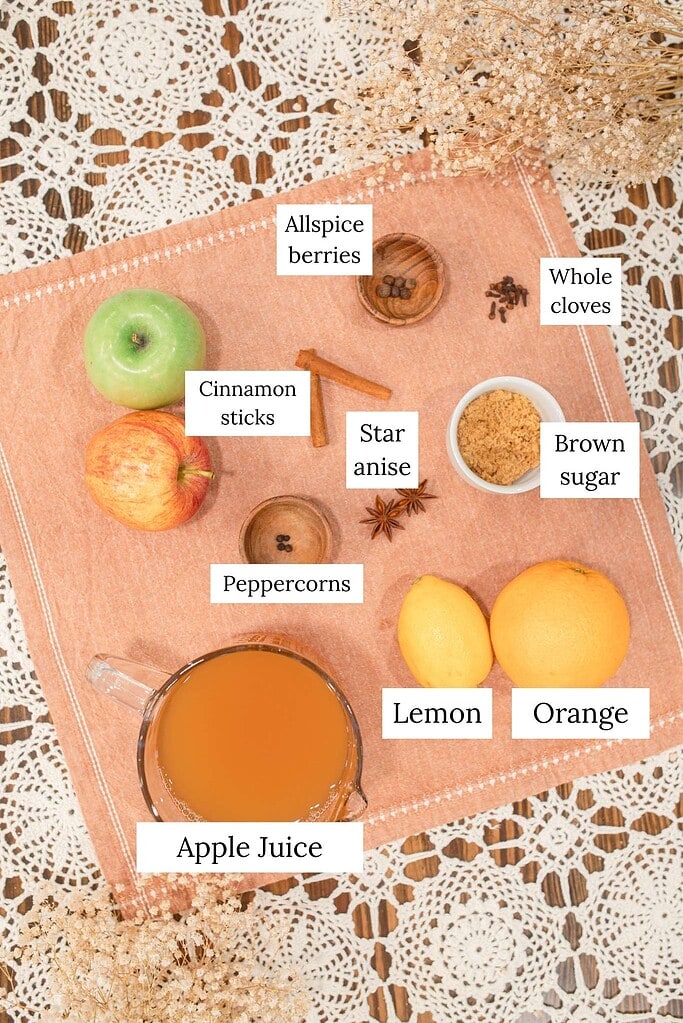 Labeled overhead ingredient flat lay for Dutch oven apple cider showing apple juice in a glass pitcher, green and red apples, allspice berries, whole cloves, cinnamon sticks, star anise, peppercorns, brown sugar, lemon, and orange arranged on a peach linen with crochet lace and dried baby's breath.