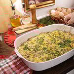 A woman in a kitchen spooning out a casserole from a 9x13" casserole white pan on wooden counter top.